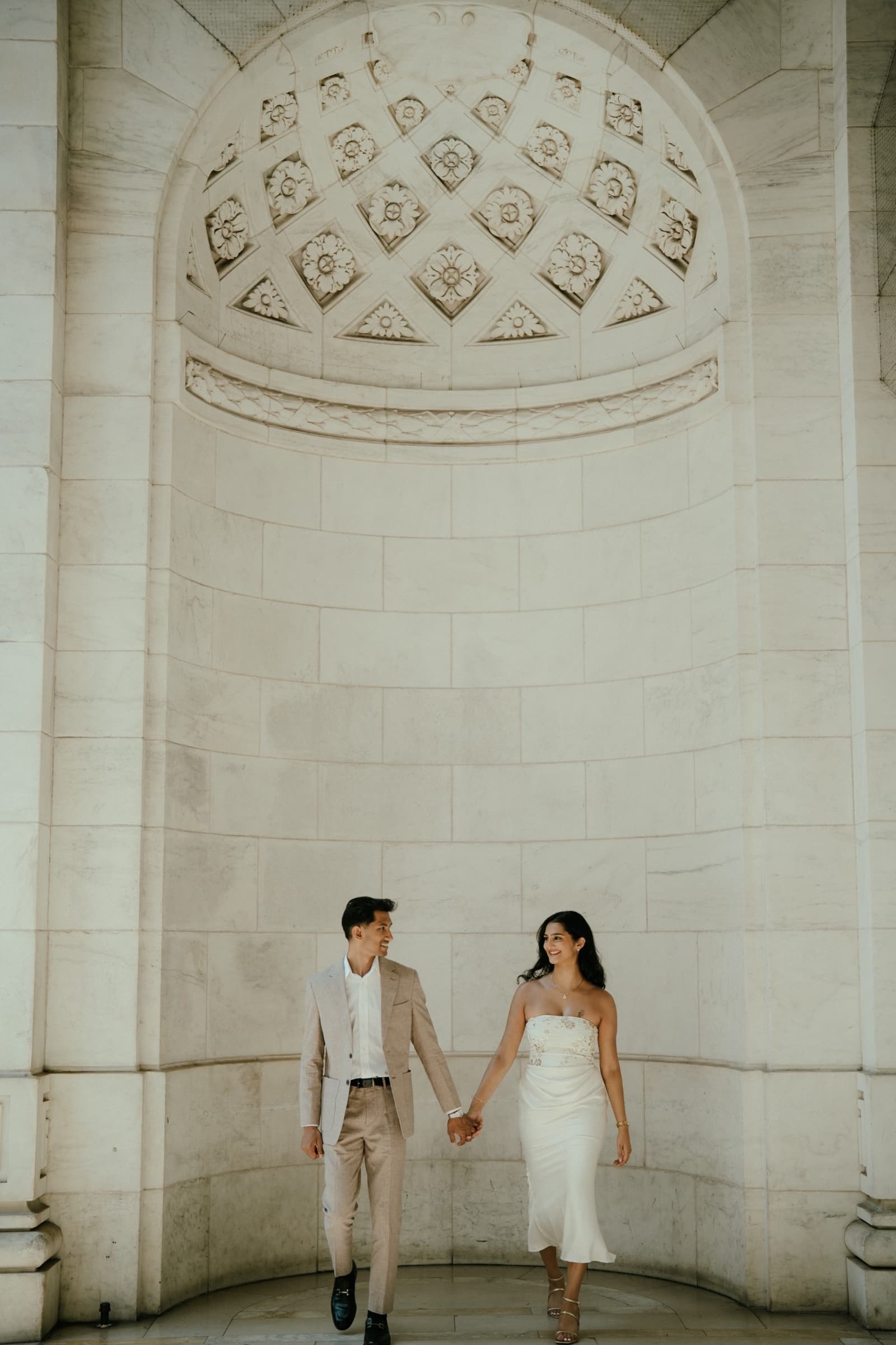 Ananya and Varun walking hand in hand beneath an ornate stone arch